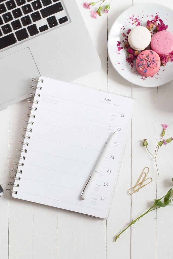 open weekly planner on top of a white desk with laptop, a plate of pink and white macarons and some flower branches
